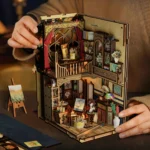 Lifestyle view of the World in a Frame book nook, featuring a two-level miniature art studio with easels, paintings, brushes, shelves, decorative furniture and warm lighting.