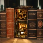 Serenity Coffee House book nook final display placed between books, featuring a miniature coffee shop scene with warm LED lighting, wooden architecture and detailed handcrafted decorations.