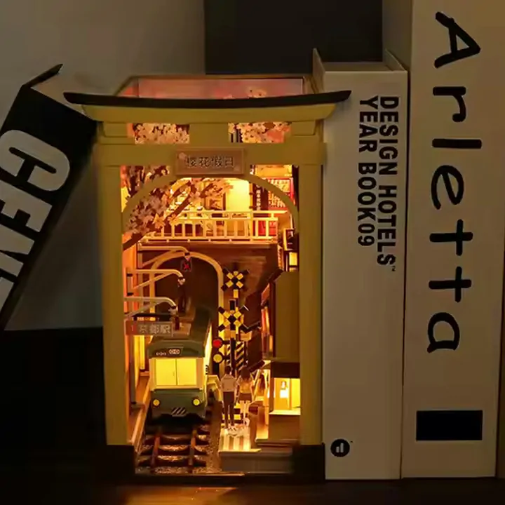 Illuminated Japan Street Cat DIY book nook displayed between books, featuring a miniature train, railway crossing, cherry blossoms, and Japanese street scenery.