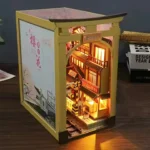 Illuminated side view of the "Japan Street Cat" DIY book nook, featuring a miniature Japanese street, shops, train tracks, and a decorative panel with cherry blossom trees.