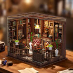 Completed and illuminated Left Bank Study DIY miniature displayed on a wooden desk, featuring tall bookshelves, leather chairs, a glowing fireplace, framed artwork, plants, and a hanging lamp.